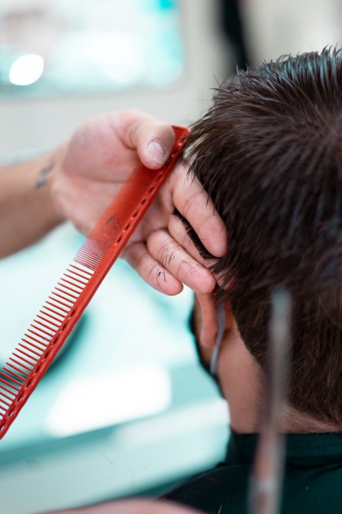 A stylist uses a red comb to shape a haircut in a barbershop.